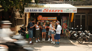 Travellers standing and eating outside Vietnamese bánh mì shop, Hoi An, Vietnam