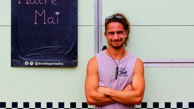 Smiling man stands beside food truck with Māori greeting sign, Muriwai, New Zealand