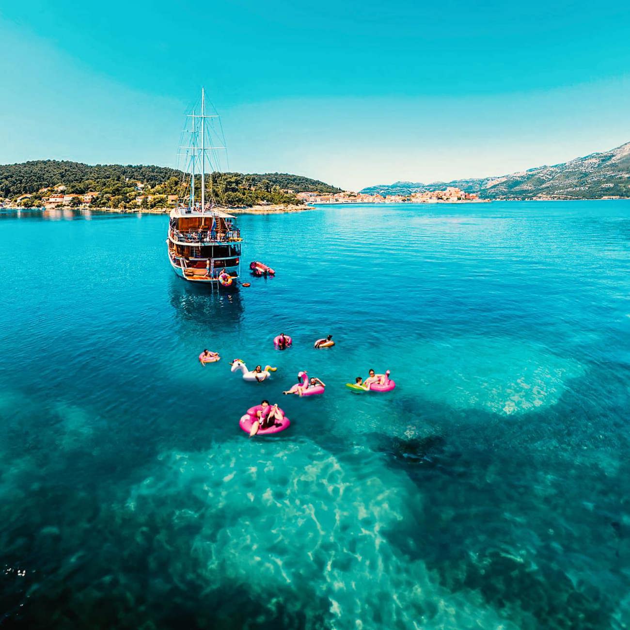 Drone view of travellers on colourful pool inflatables in clear turquoise water alongside a wooden gulet off the Croatian Dalmatian coast - Contiki Croatia Sail