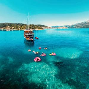 Drone view of travellers on colourful pool inflatables in clear turquoise water alongside a wooden gulet off the Croatian Dalmatian coast - Contiki Croatia Sail