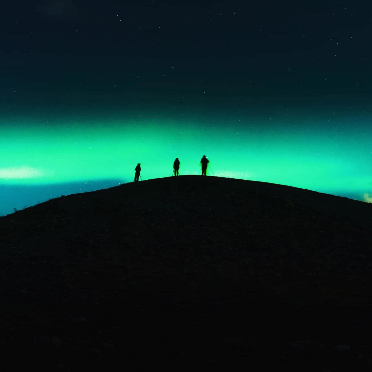 Three travellers silhouetted on a dark hilltop beneath a vivid green Aurora Borealis and starry night sky in Tromsø Norway - Contiki Norway Fjords and Northern Lights Tour
