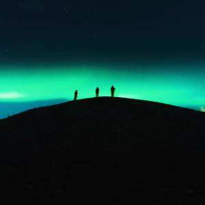 Three travellers silhouetted on a dark hilltop beneath a vivid green Aurora Borealis and starry night sky in Tromsø Norway - Contiki Norway Fjords and Northern Lights Tour