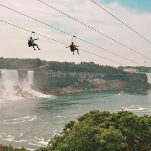 Two travellers ziplining above Niagara Falls gorge with the American and Horseshoe Falls and misty river below - Contiki Best of Canada Tour