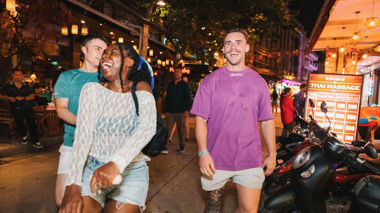Three travellers chatting and laughing, walking along a lit up street at night in Phuket, Thailand