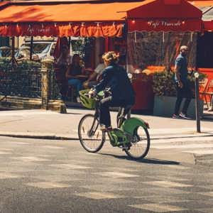 Woman cycling on a bike past a classic red-awninged French café terrace on a sunny Paris street - Contiki Europe Tour