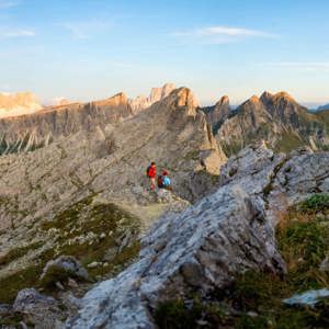 Two hikers with backpacks on a rocky mountain ridge trail at sunset with the dramatic jagged limestone peaks of the Dolomites stretching across the horizon in northern Italy - Contiki Hiking the Dolomites Tour