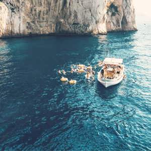 Contiki travellers swimming from a boat beneath the towering cliffs of Capri, Italy, on the Ultimate European Plus Greek Islands Tour