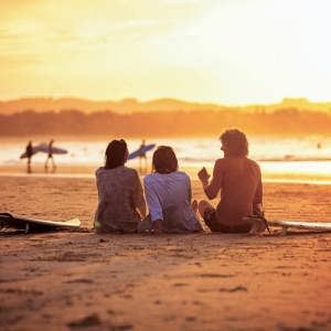 Contiki travellers relaxing on the sand with surfboards watching surfers walk along the shoreline at a stunning golden sunset in Byron Bay Australia