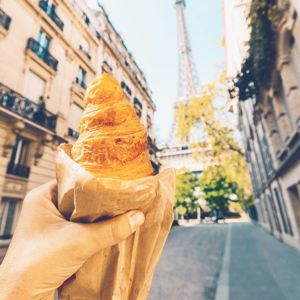 A freshly baked croissant held up towards the Eiffel Tower on a quiet Paris street during the Contiki European Horizon Tour