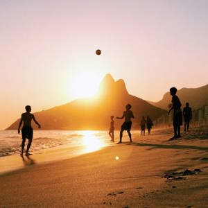 People enjoy a game of beach volleyball as the sun sets over Rio de Janeiro, with golden light reflecting on the sand and the city's dramatic mountain skyline beyond