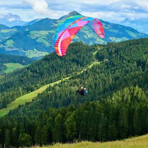 Tandem paraglider soaring over lush green pine forests and rolling Alpine meadows with a panoramic view of the Austrian Tyrol mountain valley - Contiki Active Alps Tour