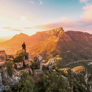 Solo traveller standing on rocky Lion's Head summit at golden hour overlooking the iconic Table Mountain and Cape Town cityscape in South Africa - Contiki Cape Town, Safari & Victoria Falls Tour