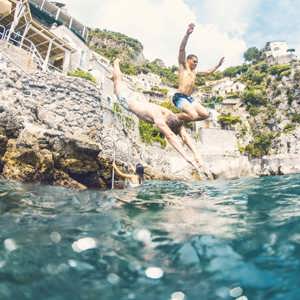 Young travellers jumping into the sea from rocks on the Amalfi Coast Italy with dramatic cliffside scenery - Contiki European Quest Tour