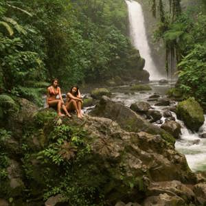 Two travellers relax on rocks beside a rainforest waterfall in Costa Rica, surrounded by lush jungle and a flowing river - Contiki Best of Costa Rica Tour