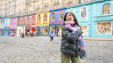 A traveller smiling standing in front of a colourful row of shops in Edinburgh, Scotland