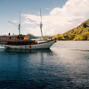 Traditional wooden phinisi sailing boat cruising through calm waters past the lush green hills of Komodo National Park Indonesia - Contiki Komodo and Flores Sailing Tour