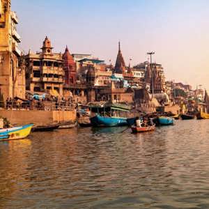 Boats drift along the Ganges River at sunrise, with the historic ghats and temples of Varanasi lining the waterfront - Delhi to Varanasi by Train Tour
