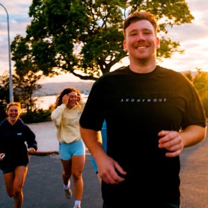 A group of travellers jog together along a lakeside road in New Zealand at sunset, smiling and enjoying the run - Contiki NZ Run Club: Muriwai Half Marathon Tour