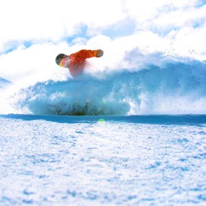 A snowboarder in a bright orange jacket rides through deep powder on a Canadian mountain, kicking up a spray of snow beneath a blue sky - Contiki Ski Canada Tour
