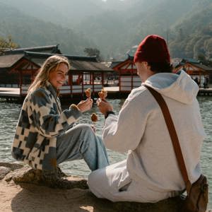Two travellers enjoy grilled street food beside the waterfront on Miyajima Island, Japan, with the iconic Itsukushima Shrine and forested mountains in the background.