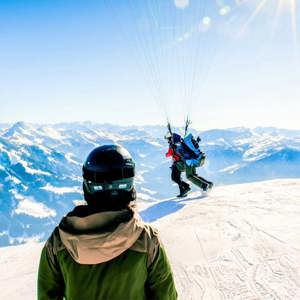 Two skiers in helmets watching a paraglider launch from a snowy Alpine summit with breathtaking panoramic views of the Austrian Alps mountain range - Contiki Ski Austria Tour