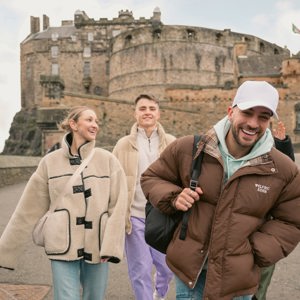 Young travellers walking in front of Edinburgh Castle in Scotland during winter, smiling and exploring the historic fortress in warm jackets on an overcast day.