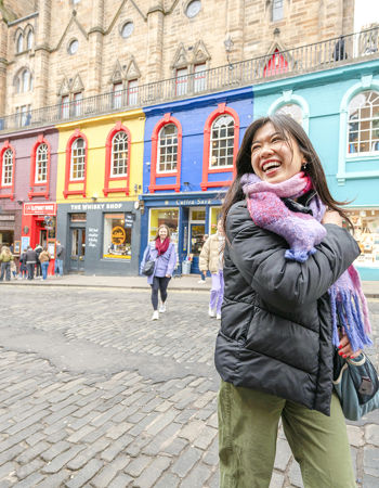 A traveller smiling standing in front of a colourful row of shops in Edinburgh, Scotland