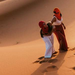 Two Contiki travellers climbing the golden sand dunes of the Sahara Desert in Morocco, dressed in traditional desert scarves during a sunset adventure on the Morocco Uncovered Tour
